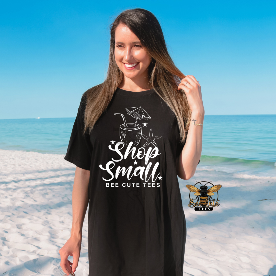 A woman stands on a sandy beach wearing a black oversized T-shirt dress with a white graphic that reads 'Shop Small' and 'BEE CUTE TEES.' The background shows blue sky and ocean.