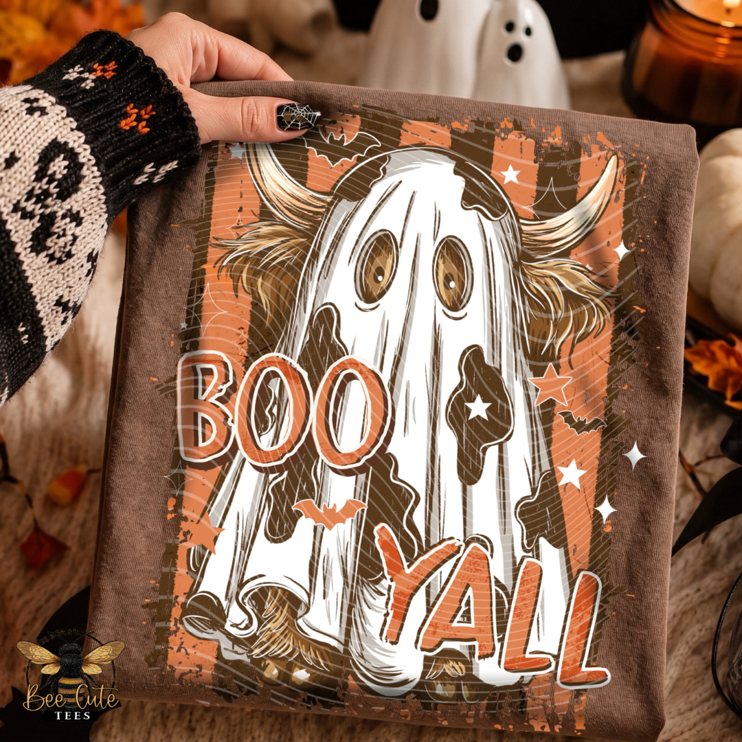 A person's hand wearing a black and white knit sweater holds up a brown fleece blanket featuring a Halloween graphic of a ghost on a bull with the text 'BOO YALL'. The background is a cozy, warm autumn setting with a white ghost decoration, a pumpkin, and a candle.