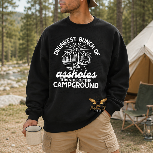A man wearing a black crewneck sweatshirt with a white graphic that reads 'DRUNKEST BUNCH OF assholes THIS SIDE OF THE CAMPGROUND' and a small bee logo reading 'Best Lads Gear' stands outdoors in a campsite setting with trees and a tent in the background.