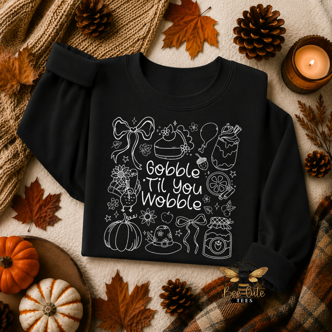 A black long-sleeve sweatshirt with a white line-art Thanksgiving-themed graphic and the phrase 'Gobble Til You Wobble' laid flat on a neutral surface with autumn props including leaves, pinecones, a candle, pumpkins, and a plaid scarf.