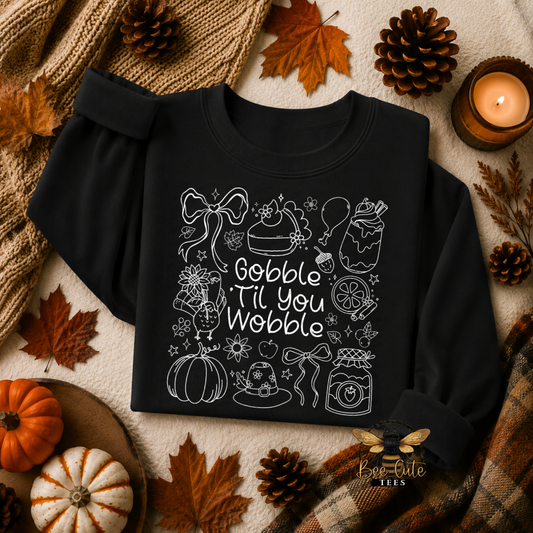 A black long-sleeve sweatshirt with a white line-art Thanksgiving-themed graphic and the phrase 'Gobble Til You Wobble' laid flat on a neutral surface with autumn props including leaves, pinecones, a candle, pumpkins, and a plaid scarf.