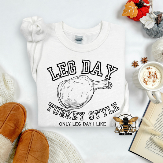 A white graphic T-shirt laid flat on a light background with cozy props. The shirt has black text reading 'LEG DAY TURKEY STYLE' and 'ONLY LEG DAY I LIKE' around a turkey leg illustration.