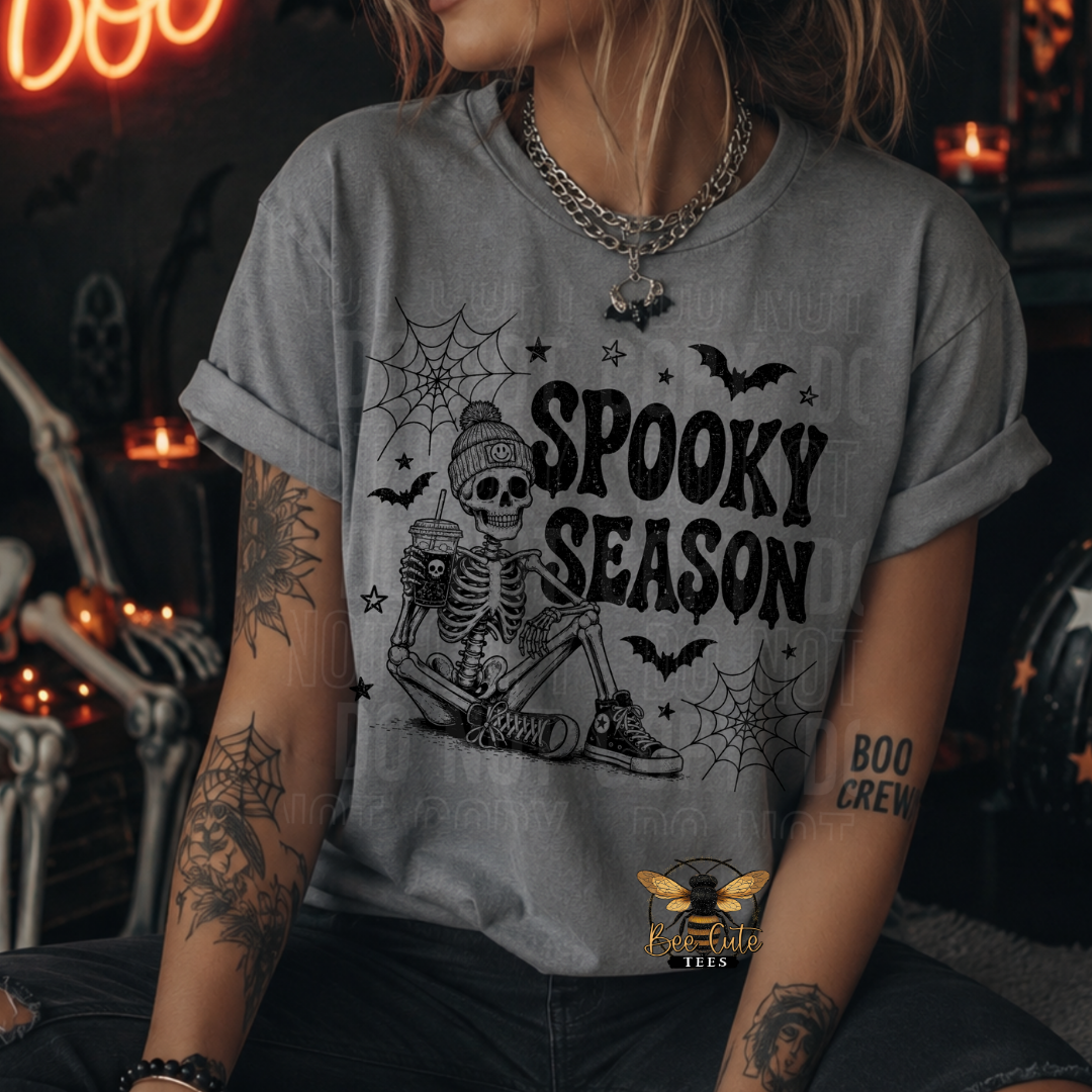 A woman wearing a heather gray graphic t-shirt with a black 'Spooky Season' skeleton design, posed in a dark, cozy Halloween-themed room with candles and neon lights.