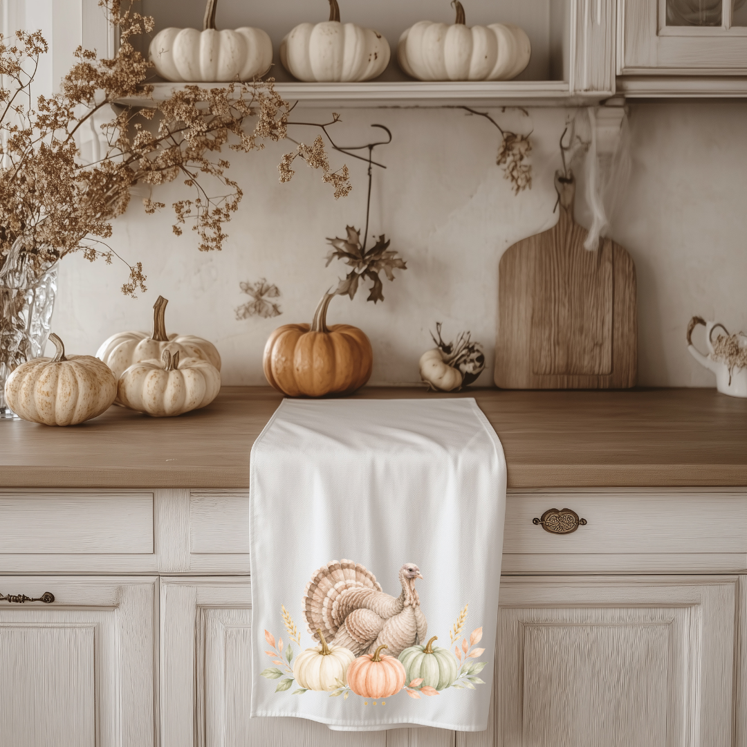 A white tea towel with a fall turkey and pumpkins print draped over a kitchen counter, surrounded by autumn decorations like pumpkins, a wooden cutting board, and dried flowers.