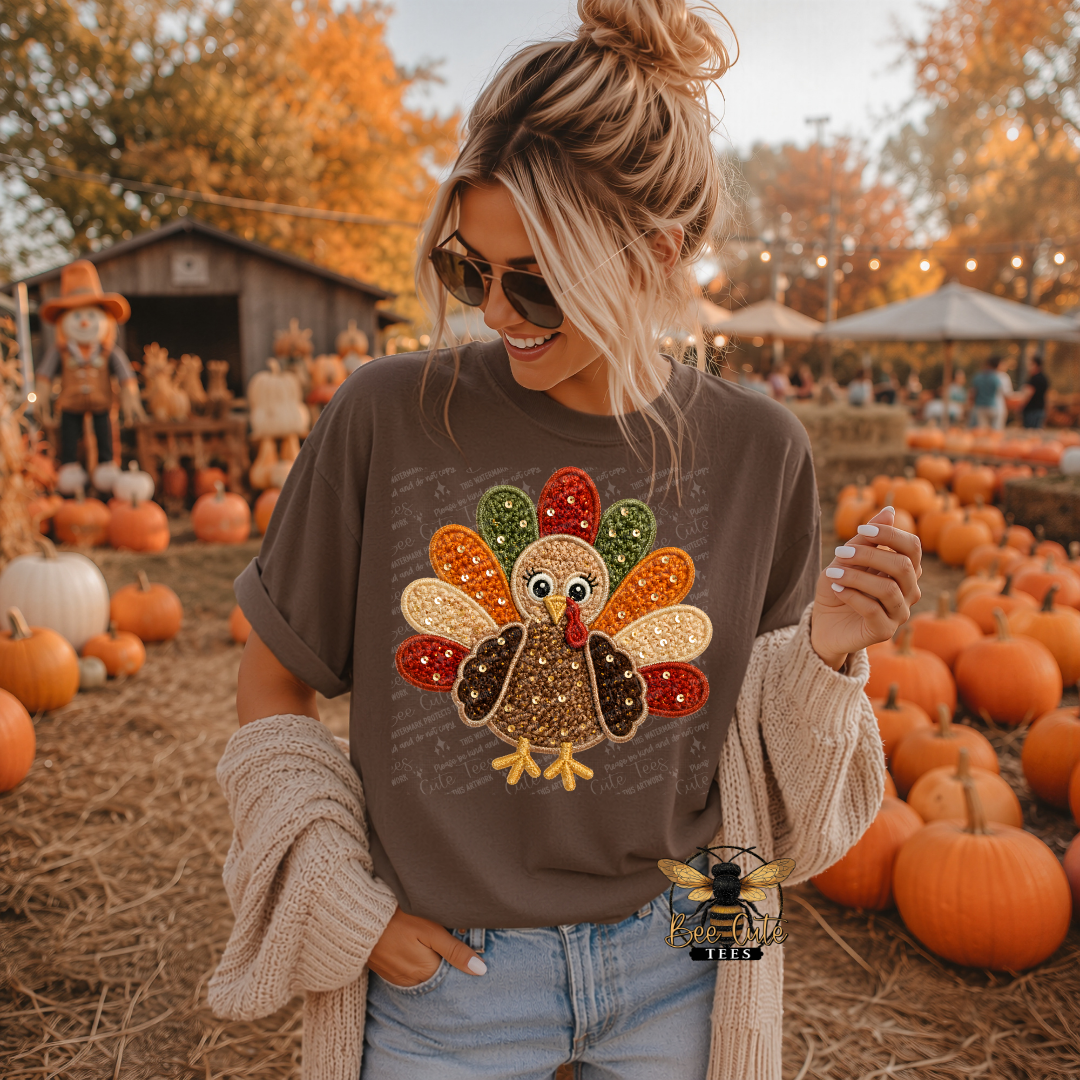 A woman wearing a brown graphic t-shirt with a large, colorful, textured turkey applique on the front, standing in a pumpkin patch with a beige knit cardigan draped off her shoulders.