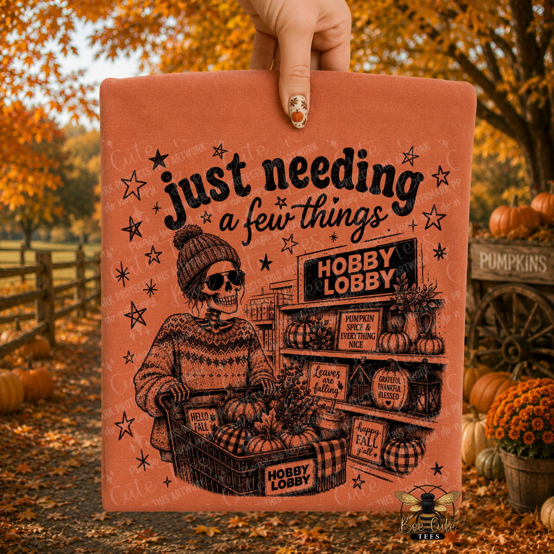 A hand holds up an orange folded t-shirt featuring a skeleton shopper at a store with pumpkins and autumn decor, set against an outdoor fall yard scene with pumpkins and orange foliage.