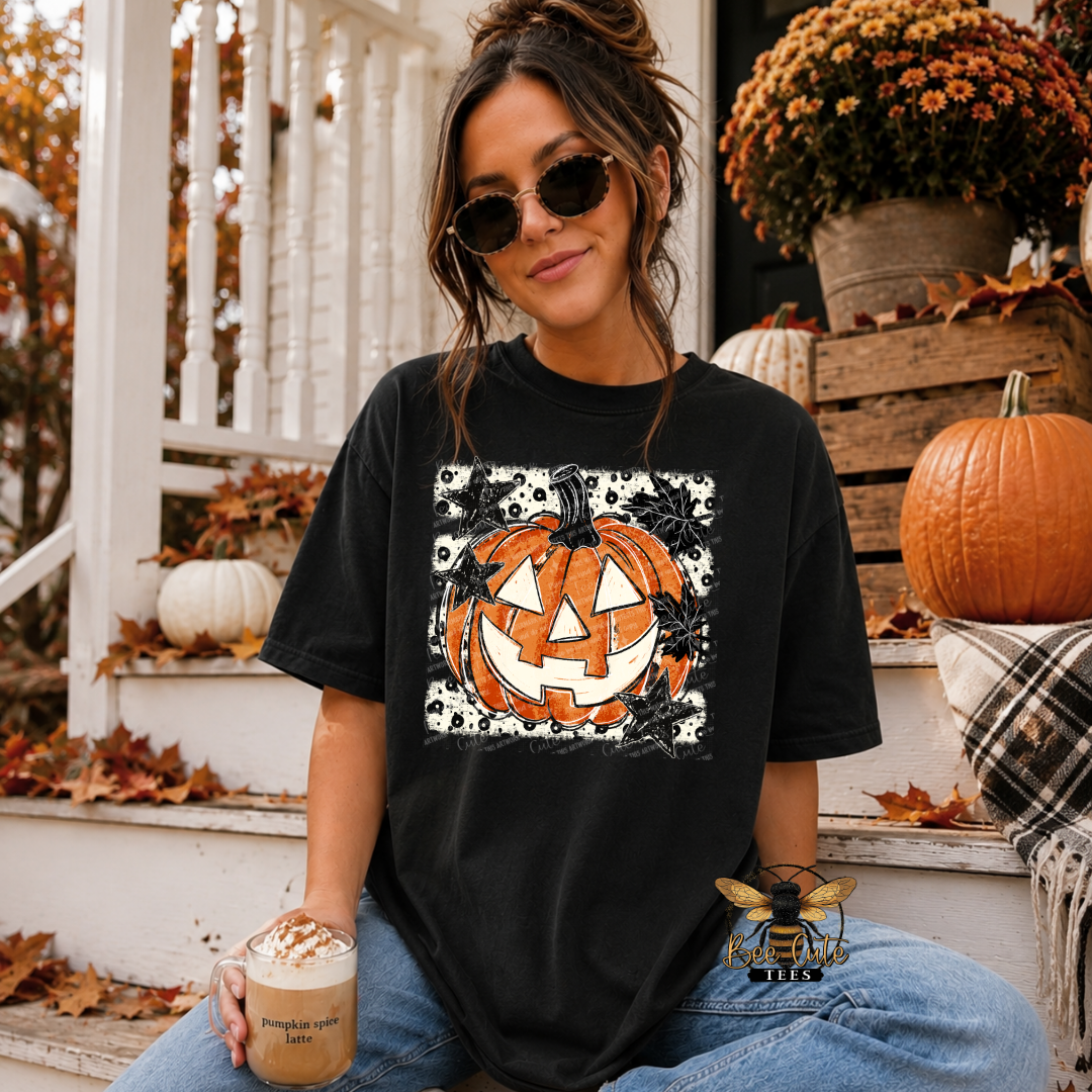 A woman wearing a black graphic t-shirt with a jack-o'-lantern design, sitting on a porch decorated for autumn with pumpkins and fall leaves.