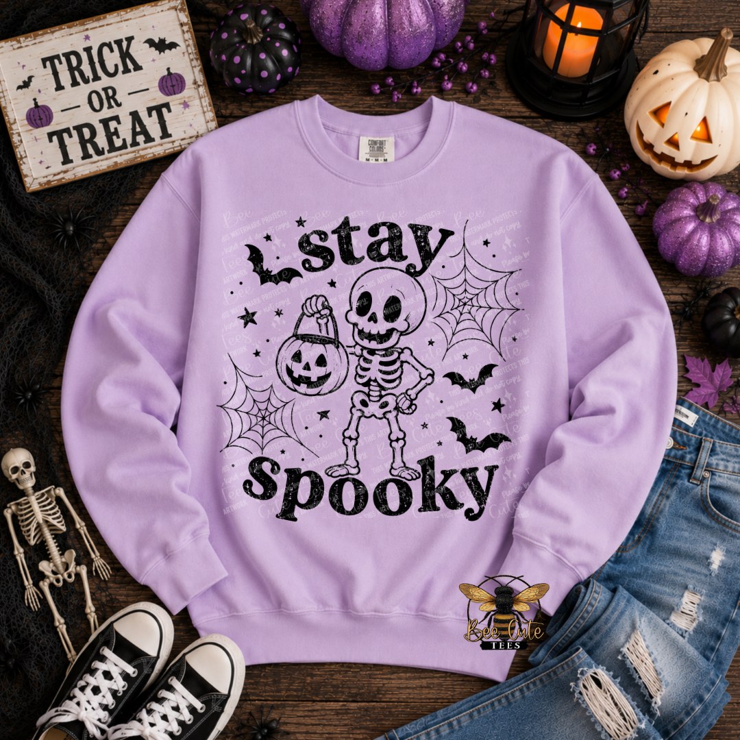 A lavender crewneck sweatshirt with a black 'stay spooky' skeleton and jack-o'-lantern graphic, styled in a flat lay with Halloween props including pumpkins, a lantern, jeans, sneakers, and a small skeleton figurine.