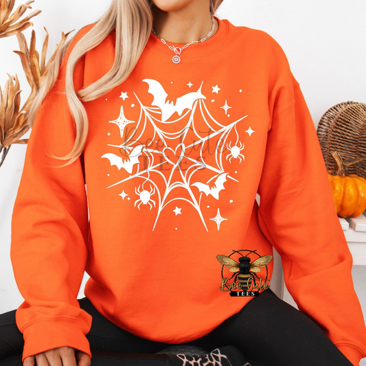 A woman wearing a bright orange sweatshirt with a white spiderweb, bats, spiders, and stars graphic on the front. The photo is cropped from the chin down, with decorative dried grass on the left and pumpkins on the right.