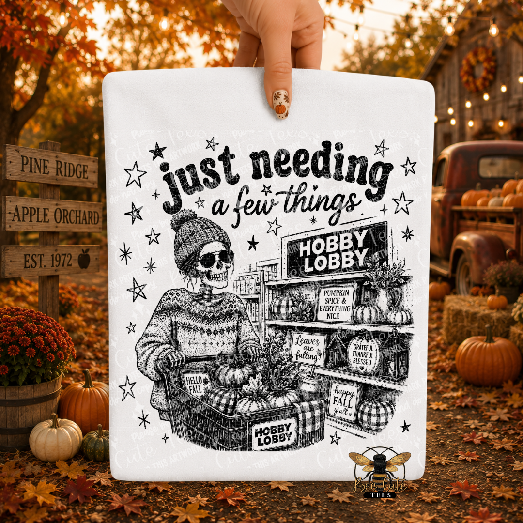 A white folded t-shirt held by a hand, featuring a black illustrated skeleton in a beanie and sweater pushing a cart in front of autumn-themed fall foliage, pumpkins, and rustic signs. The sign reads “just needing a few things”.