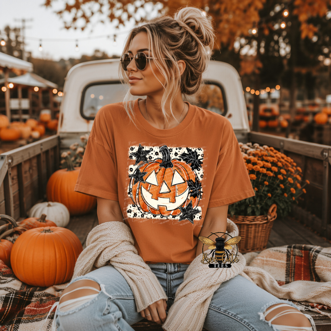 A woman wearing a burnt orange graphic t-shirt with a jack-o'-lantern design, sitting in the bed of a pickup truck surrounded by pumpkins and fall decorations.