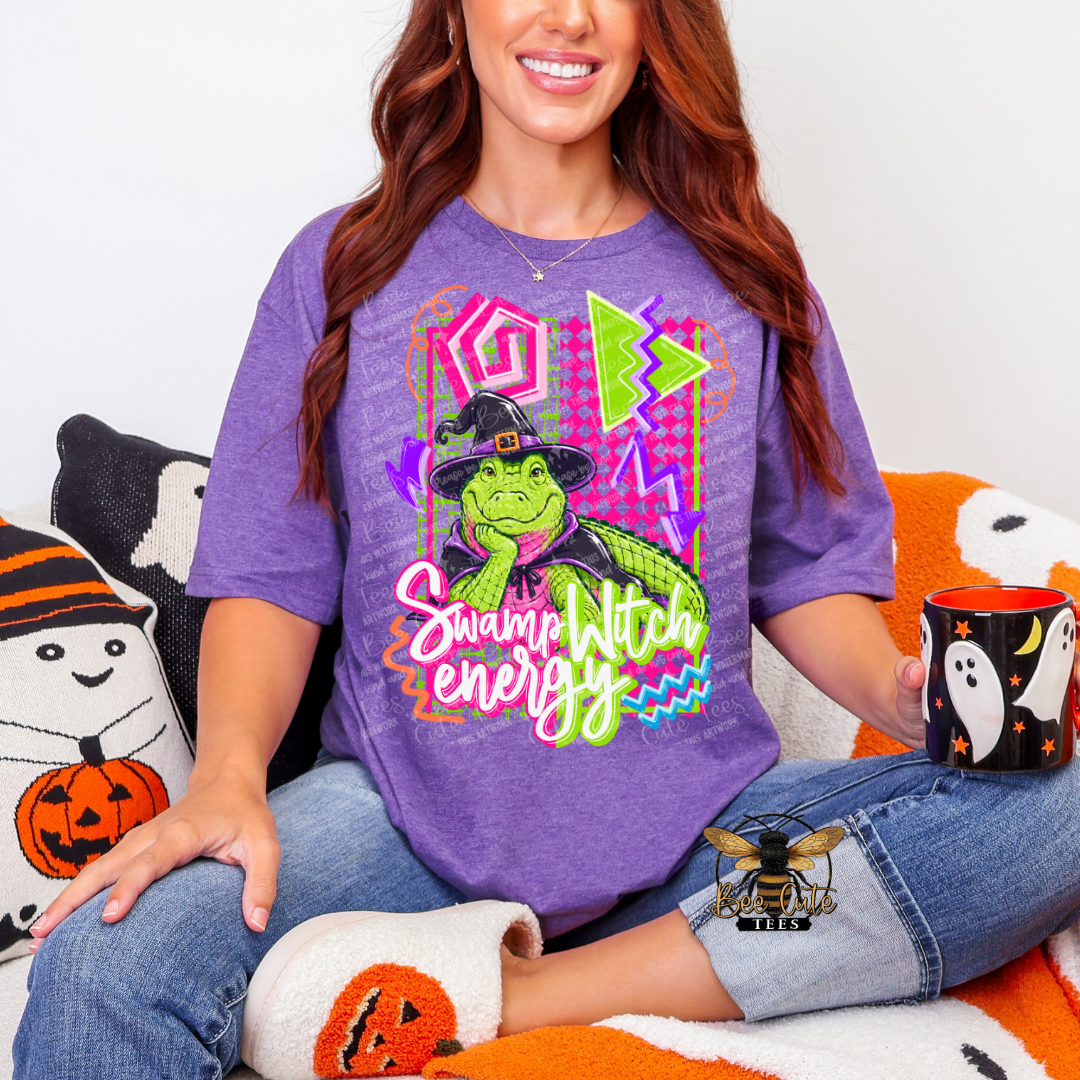 A woman wearing a purple graphic t-shirt with a green witch character and the text 'Swamp Witch energy' in neon colors, sitting on a Halloween-themed couch and holding a black mug with a ghost design.