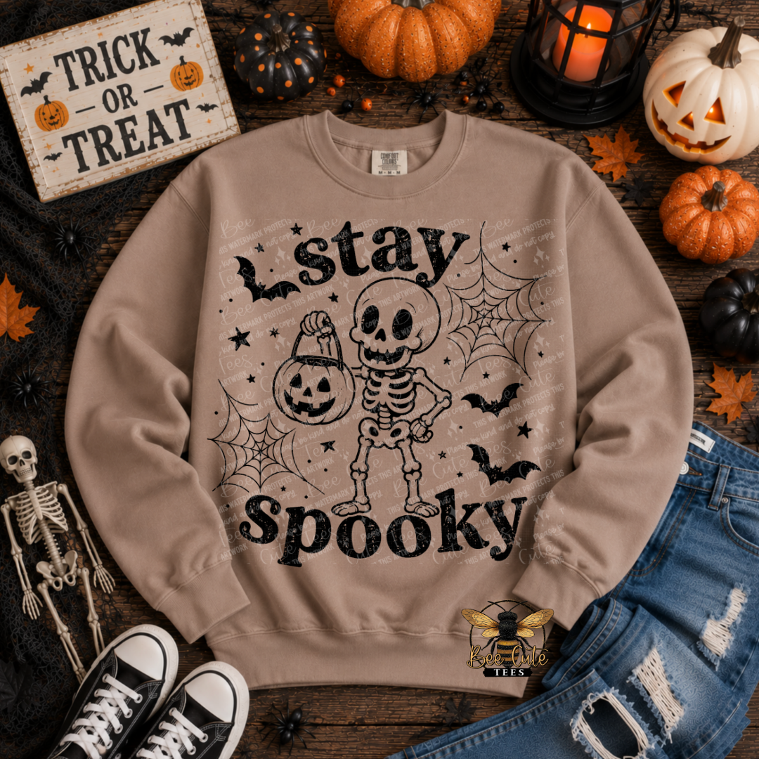A beige crewneck sweatshirt with a distressed black graphic of a skeleton holding a jack-o'-lantern, surrounded by spooky elements like pumpkins, a sign reading 'Trick or Treat', jeans, sneakers, and a small bee logo, arranged in a flat lay.