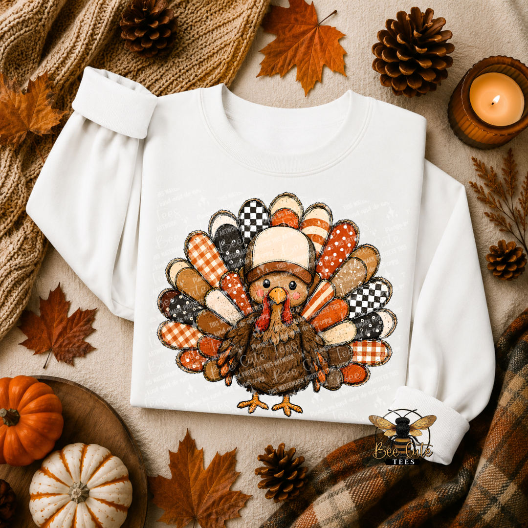 A white long-sleeve sweatshirt laid flat on a cozy autumn-themed background, featuring a large embroidered-style turkey graphic in orange, brown, beige, black, and white patterned feathers. The scene includes fall leaves, pinecones, pumpkins, a knitted scarf, and a lit candle.