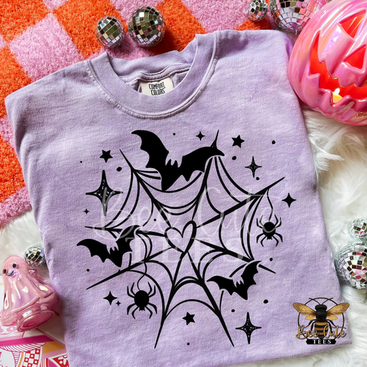 A lavender crewneck t-shirt with a black spiderweb, bats, spiders, and stars graphic, styled in a flat lay with Halloween props including a jack-o'-lantern, mini disco balls, and a pink ghost figure.