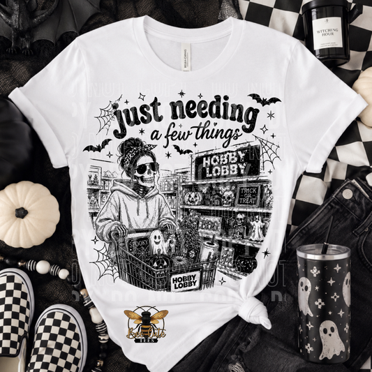 A white graphic t-shirt featuring a black and white illustration of a skeleton shopper in a Halloween-themed store, styled in a flat-lay with dark denim, checkered shoes, a pumpkin, and a tumbler.
