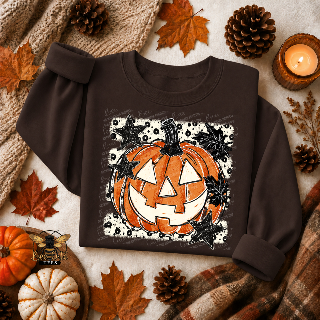 A dark brown crewneck sweatshirt with a Halloween graphic of a smiling jack-o'-lantern surrounded by black leaves on a white splattered background. The sweatshirt is styled in a flat lay with autumn props including pinecones, maple leaves, a small pumpkin, a knitted blanket, and a lit candle.