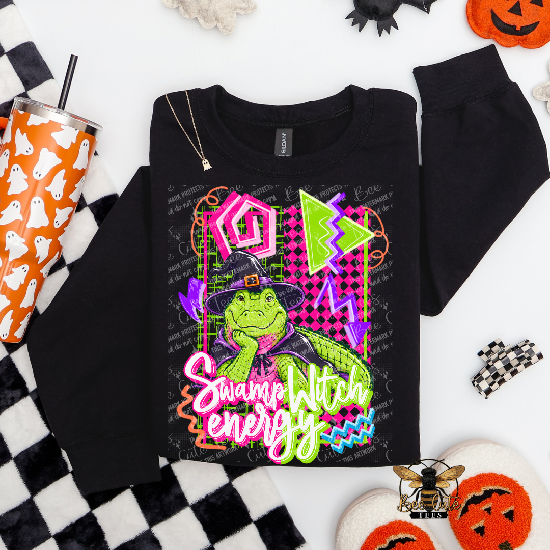 A black long-sleeve sweatshirt with a colorful Swamp Witch graphic, styled in a flat lay with Halloween props including a ghost-patterned tumbler, black-and-white checkered fabric, and orange pumpkins on a white background.