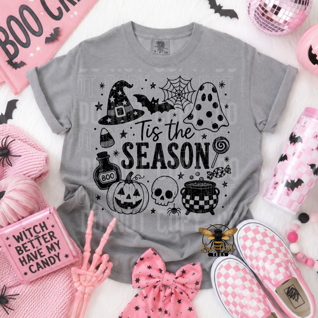 A gray short-sleeve graphic T-shirt laid flat on a white textured surface, surrounded by pink and black Halloween-themed props including a pink bow, shoes, a tumbler, and candy-themed accessories. The shirt graphic reads 'Tis the Season' with witch hats, bats, a spiderweb, a pumpkin, skull, cauldron, and other spooky motifs.