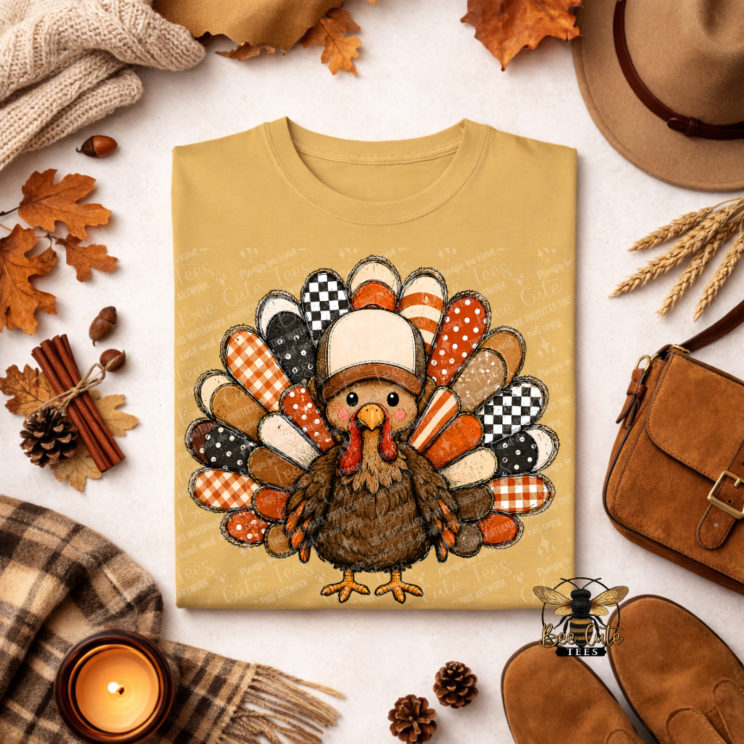 A folded mustard yellow T-shirt with a large Thanksgiving turkey graphic on the front, styled in a flat lay with autumn leaves, a hat, shoes, a candle, and a small bee logo near the bottom right.
