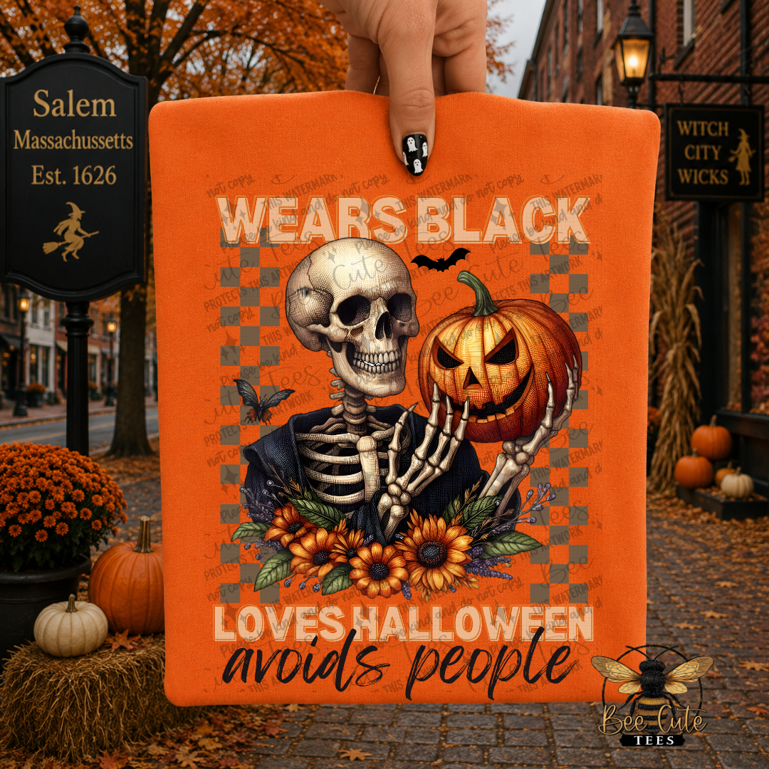 An orange rectangular graphic print held by a hand, featuring a Halloween design with a skeleton, jack-o'-lantern, sunflowers, and the text 'WEARS BLACK LOVES HALLOWEEN avoids people'. The background is a blurred autumn street scene with Halloween decorations.