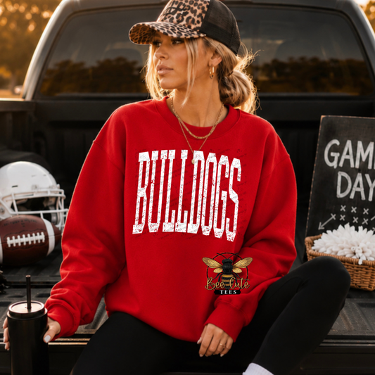 A woman wearing a red crewneck sweatshirt with 'BULLDOGS' printed in distressed white letters across the chest, sitting in the bed of a truck. She is wearing a leopard-print cap and black pants, with football gear and a 'Game Day' sign in the background.