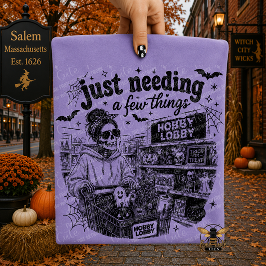 A hand holds a square purple folded t-shirt featuring a black-and-white illustrated skeleton in a store setting with the text 'Just needing a few things.' The background is a Halloween-themed street scene with autumn leaves, pumpkins, and signs reading Salem, Massachusetts. 