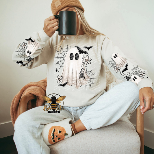 A woman sitting in a chair wearing a cream-colored sweatshirt with a large ghost and spiderweb Halloween graphic on the front, paired with light gray sweatpants and a brown beanie while holding a black mug.
