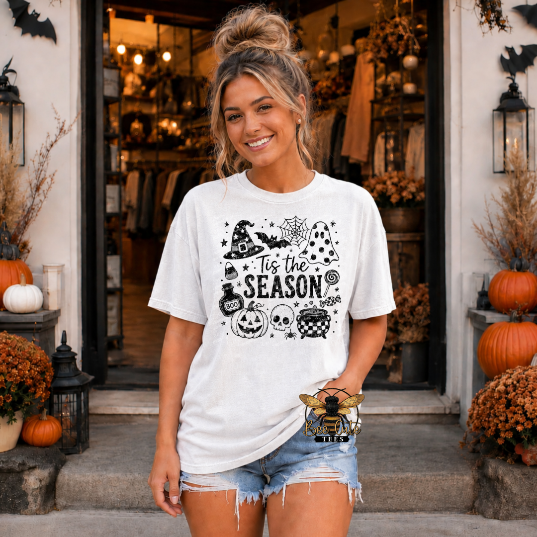 A woman stands in front of a storefront doorway wearing a white graphic T-shirt with black Halloween-themed illustrations and the text 'Tis the Season.' She is also wearing denim shorts, and the setting includes pumpkins, lanterns, and fall decorations.