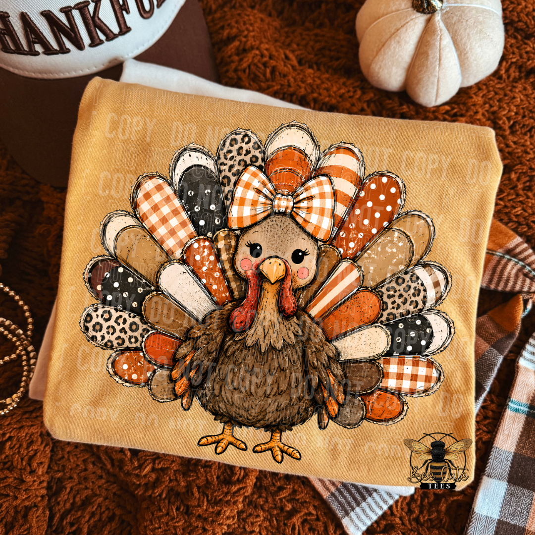 A folded mustard yellow t-shirt  featuring a cartoon turkey surrounded by patterned feathers in autumn colors, placed on a brown textured blanket with seasonal props.