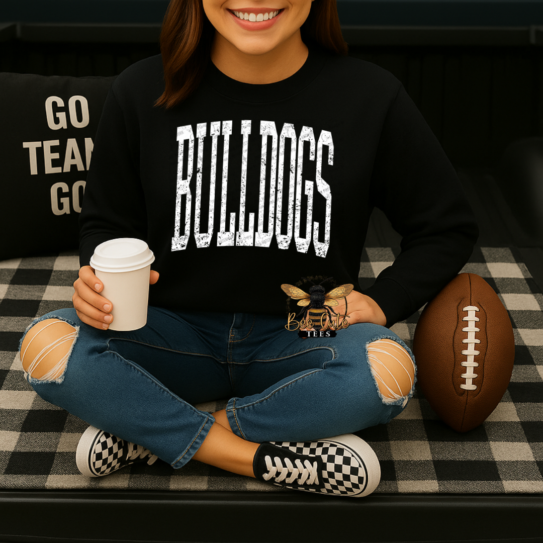 A woman sitting cross-legged on a black-and-white checkered couch, wearing a black crewneck sweatshirt with 'BULLDOGS' printed in distressed white block letters across the chest. She is holding a white coffee cup and wearing ripped blue jeans and black-and-white sneakers.