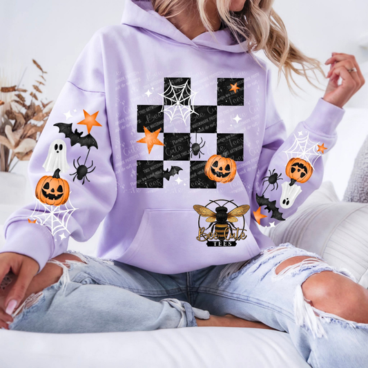 A woman wearing a light purple hoodie with a Halloween-themed graphic print featuring a checkerboard pattern, pumpkins, ghosts, spiders, and a bee, paired with ripped light blue jeans.