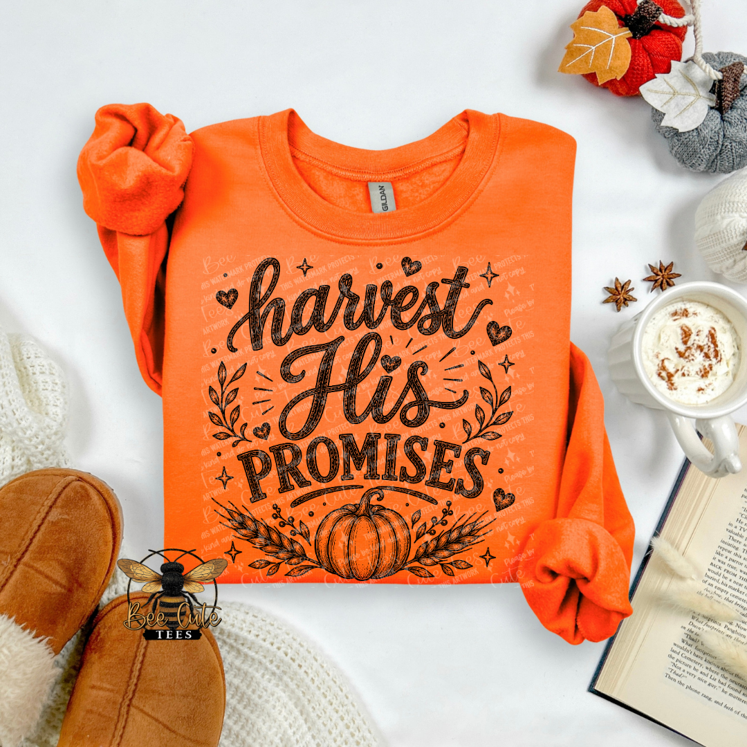 An orange crewneck sweatshirt with a dark brown graphic reading 'harvest His PROMISES' and a pumpkin design, styled in a cozy flat-lay with a mug of hot chocolate, an open book, and autumn-themed props on a white background.