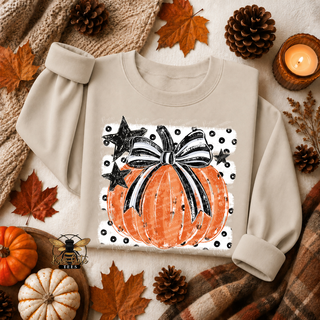 A beige long-sleeve sweatshirt laid flat with a large autumn-themed pumpkin graphic on the chest. The design shows an orange pumpkin with a black and white bow, black stars, and a white polka-dot border.