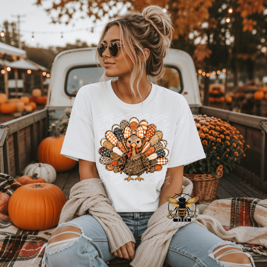 A woman sitting in the bed of a pickup truck wearing a white graphic t-shirt with a colorful autumn turkey design. The scene is decorated with pumpkins, fall foliage, and cozy blankets.