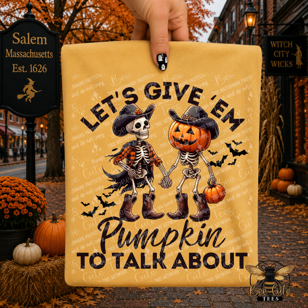 A hand holds a mustard yellow folded t-shirt, featuring a Halloween graphic of two skeletons holding hands with pumpkins and the text 'LET'S GIVE 'EM Pumpkin TO TALK ABOUT.' The background is a blurred autumn street scene with pumpkins, fall leaves, and storefront signs, including Salem Massachusetts and Witch City Wicks.