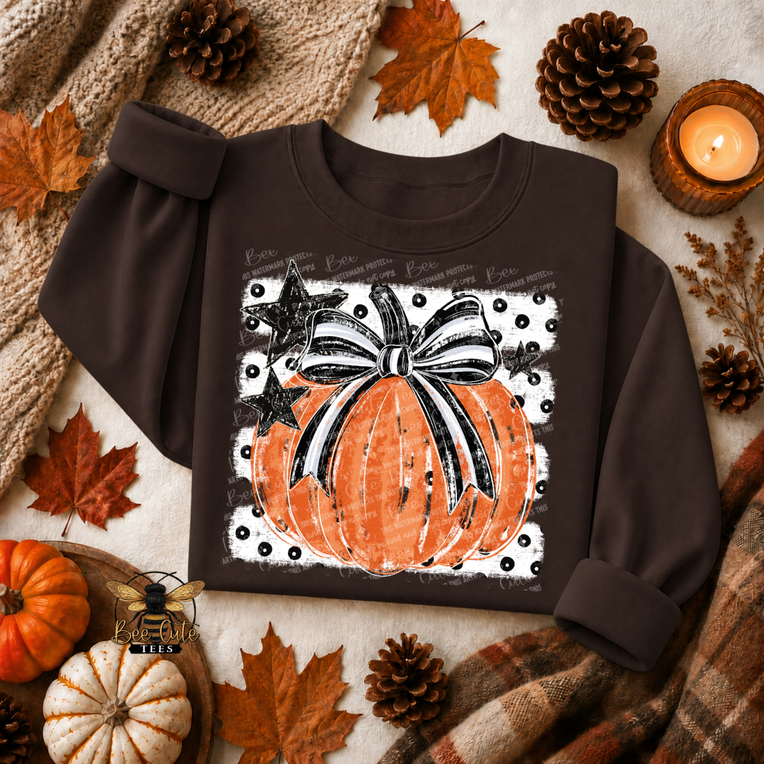 A dark brown long-sleeve sweatshirt laid flat on a cozy autumn-themed background. The sweatshirt features a graphic print of an orange pumpkin with a black and white striped bow, set against a white background with black polka dots and a black star.
