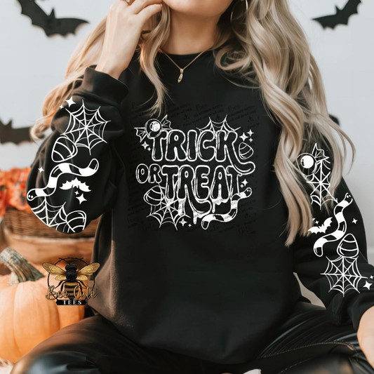 A woman wearing a black crewneck sweatshirt with a white 'Trick or Treat' graphic and spiderwebs printed on both sleeves, posed with pumpkins and Halloween decorations in the background.