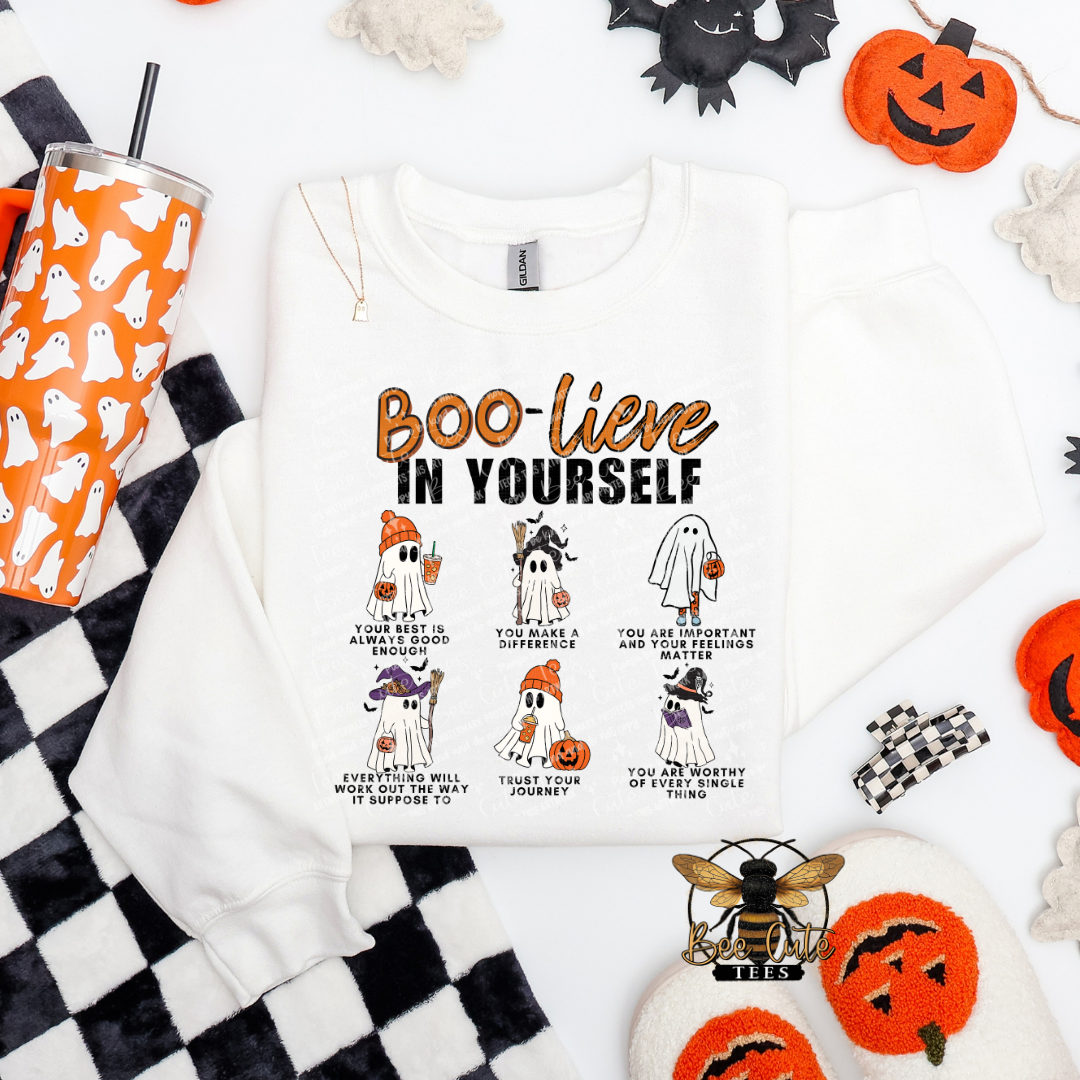 A white crewneck sweatshirt laid flat on a white background with Halloween-themed props. The sweatshirt features a graphic print that reads 'BOO-lieve IN YOURSELF' above six small illustrated ghosts in costumes with encouraging phrases.