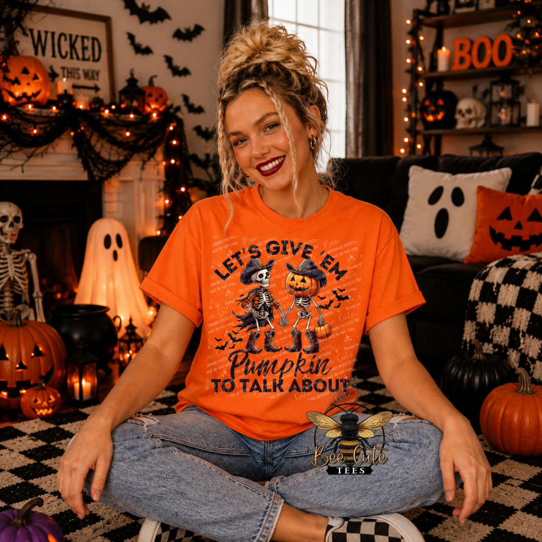 A woman sitting cross-legged in a Halloween-themed living room wearing an orange graphic t-shirt that reads 'LET'S GIVE 'EM Pumpkin TO Talk ABOUT' with cartoon skeletons and pumpkins. The background features a decorated fireplace, jack-o'-lanterns, a ghost lamp, and a checkered rug.