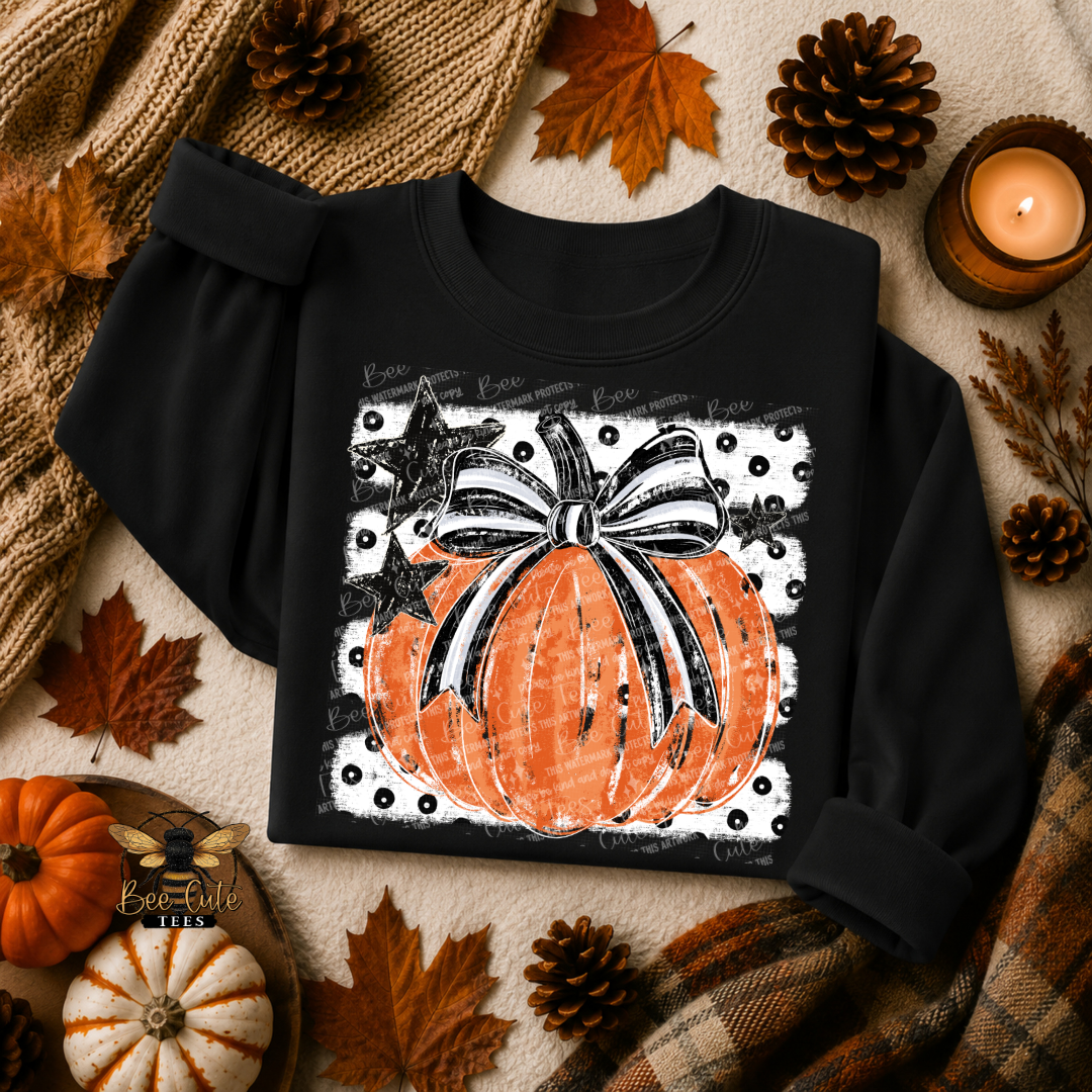 A black long-sleeve sweatshirt laid flat on a cozy autumn-themed background with a printed pumpkin graphic featuring an orange pumpkin, black and white striped bow, white polka dot border, and handwritten-style text. The scene includes pumpkins, pinecones, leaves, a candle, and plaid fabric.