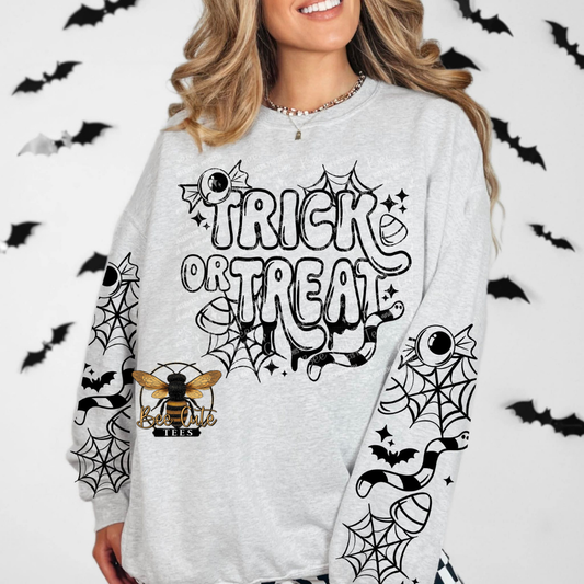 A woman wearing a light gray long-sleeve sweatshirt with a black and white 'Trick or Treat' spiderweb graphic on the front, decorated with spiderwebs, bats, and a bee on the sleeves, set against a white background with black bat silhouettes.