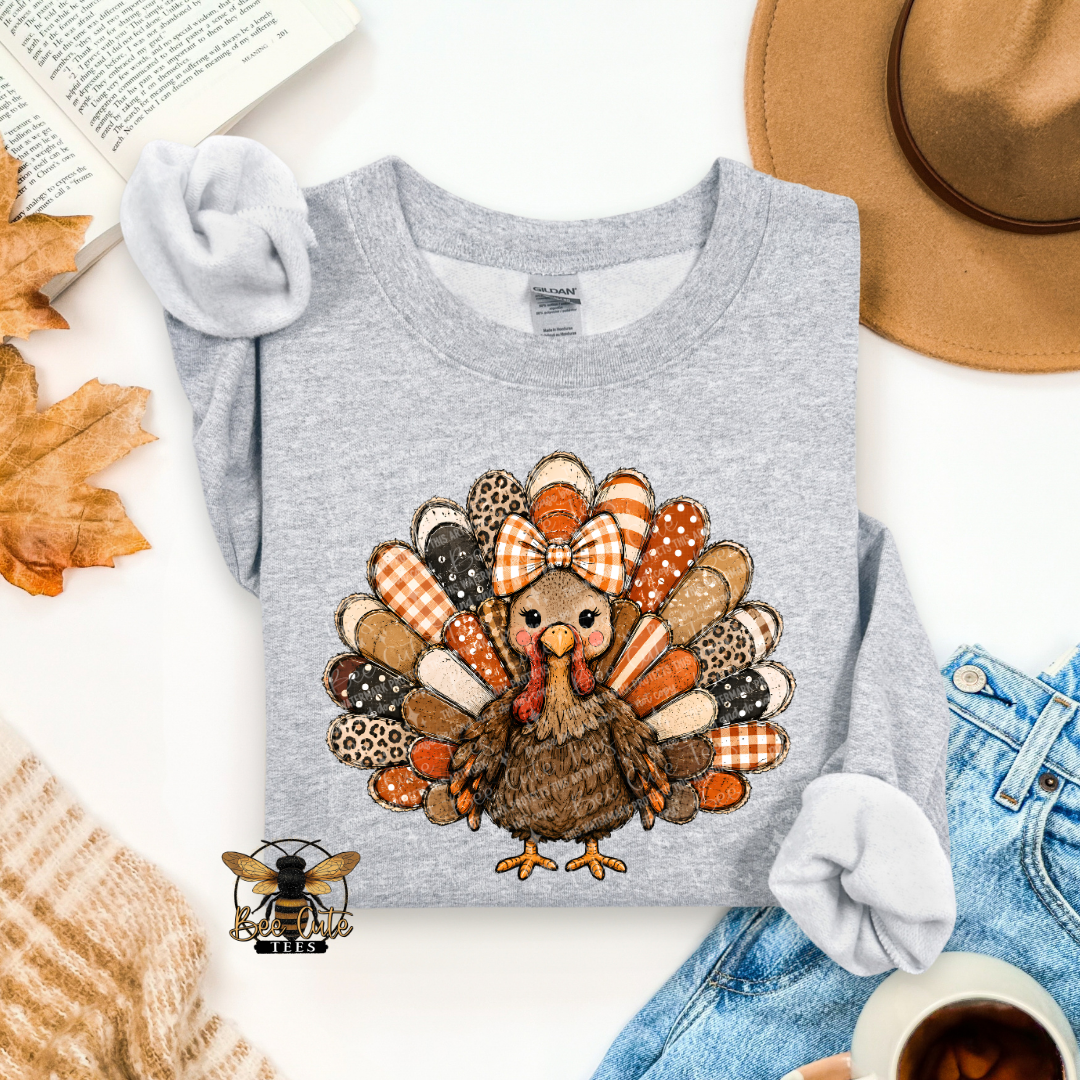 A folded heather gray long-sleeve t-shirt with a colorful Thanksgiving turkey graphic on the front, styled in a flat lay with a tan hat, blue jeans, a knit sweater, an open book, autumn leaves, and a cup of coffee.