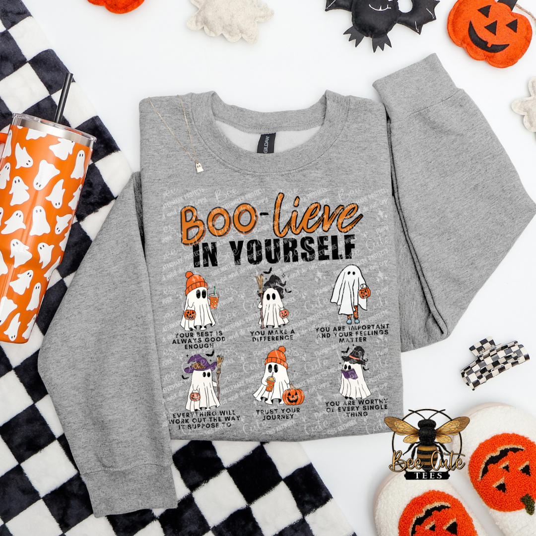 A heather gray crewneck sweatshirt with a Halloween-themed graphic print reading 'BOO-lieve IN YOURSELF' above six cartoon ghosts in different outfits and poses. The sweatshirt is styled in a flat lay with Halloween props including a black-and-white checkered blanket, an orange cup, jack-o'-lanterns, and a bee logo watermark.