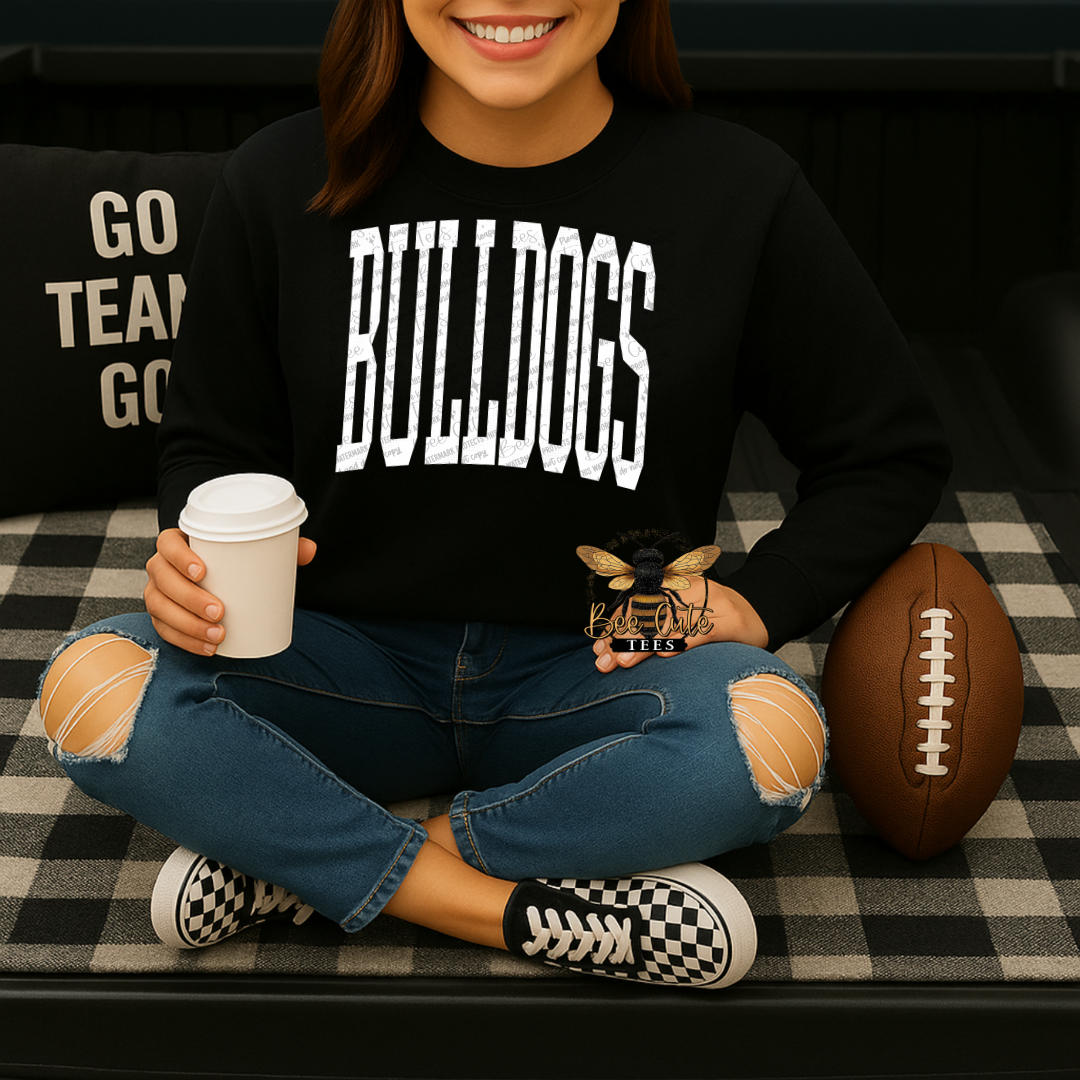 A woman sitting cross-legged on a black-and-white checkered couch, wearing a black sweatshirt with 'BULLDOGS' printed in large white letters across the chest. She is holding a coffee cup and a small bee figurine, with a football beside her and a pillow reading 'GO TEAM GO' behind her.