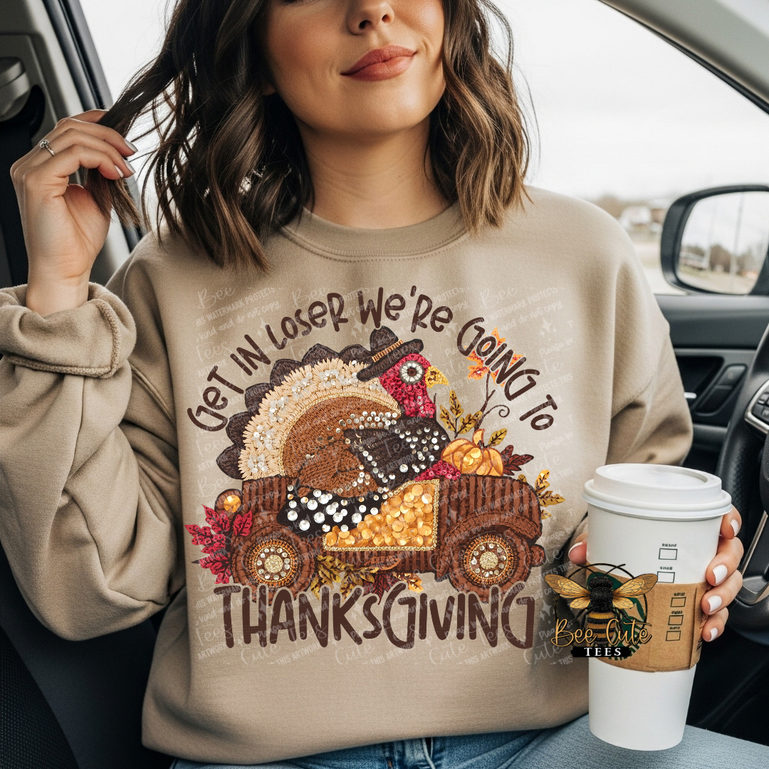 A woman sitting inside a car wearing a beige crewneck sweatshirt with a colorful Thanksgiving graphic of a turkey in a truck and the text 'GET IN LOSER WE'RE GOING TO THANKSGIVING'. She is holding a white coffee cup.
