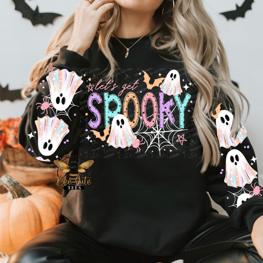 A woman wearing a black long-sleeve sweatshirt with a colorful Halloween graphic that reads 'let's get Spooky' in pastel letters, decorated with cartoon ghosts, spiders, bats, and spiderwebs. She is sitting in a cozy setting with pumpkins and black bat decorations in the background.