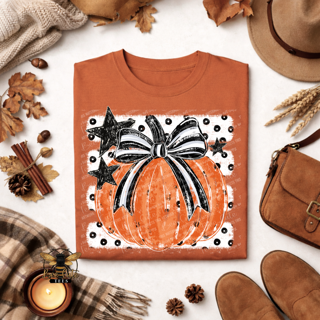 An orange crewneck t-shirt with a distressed graphic of a pumpkin, black stars, and a black-and-white striped bow, styled in an autumn flat lay with a hat, scarf, candle, and boots.