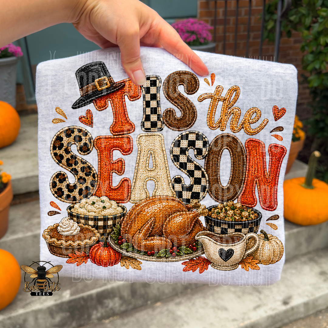 A hand holds a white fabric cross-stitch sampler featuring the phrase 'THIS IS THE SEASON' in decorative autumn-themed letters, with a turkey, pie, pumpkin, and gravy motif below. The background shows outdoor steps with pumpkins and plants.