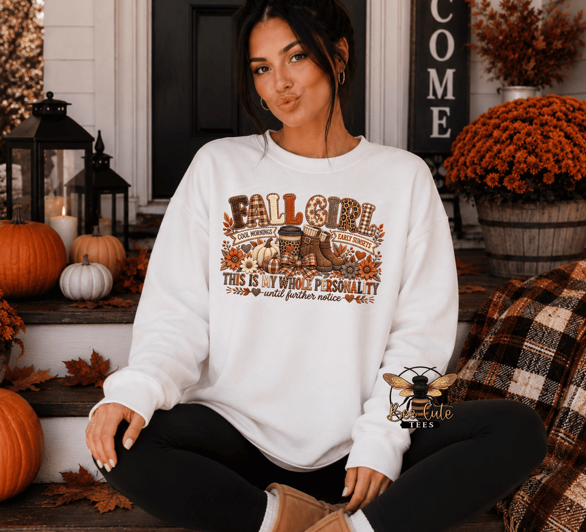 A woman sits cross-legged on a decorated front porch wearing a white crewneck sweatshirt with a brown and orange 'Fall Girl' graphic. The sweatshirt features a design with pumpkins, a turkey, and autumn-themed text.
